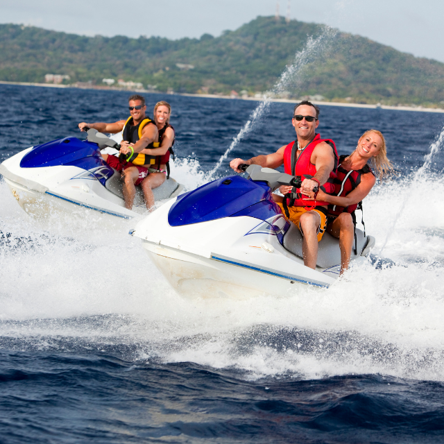 Two people on each of two jet skis, riding on blue water, smiling, wearing life vests. Lush green hills in the background.
