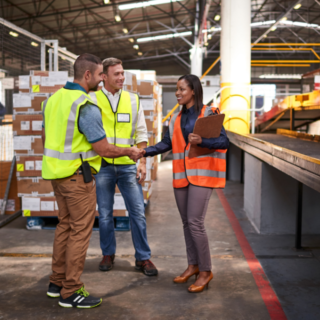 Three people in a warehouse shake hands; all wearing safety vests. Boxes and conveyor belt in the background.