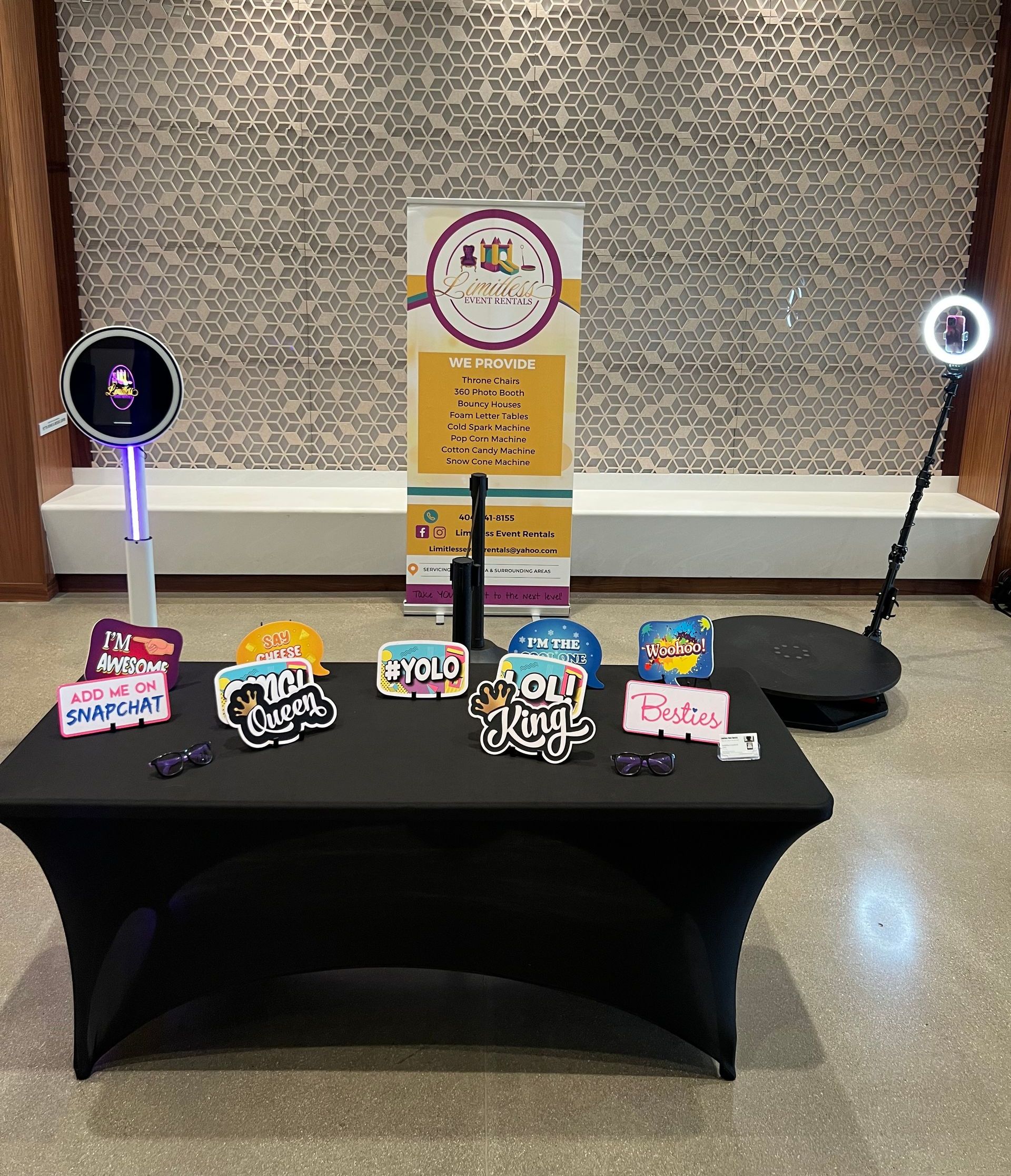 Photo booth setup with props on a black table: a sign, ring light, and photo booth machine against a textured wall.