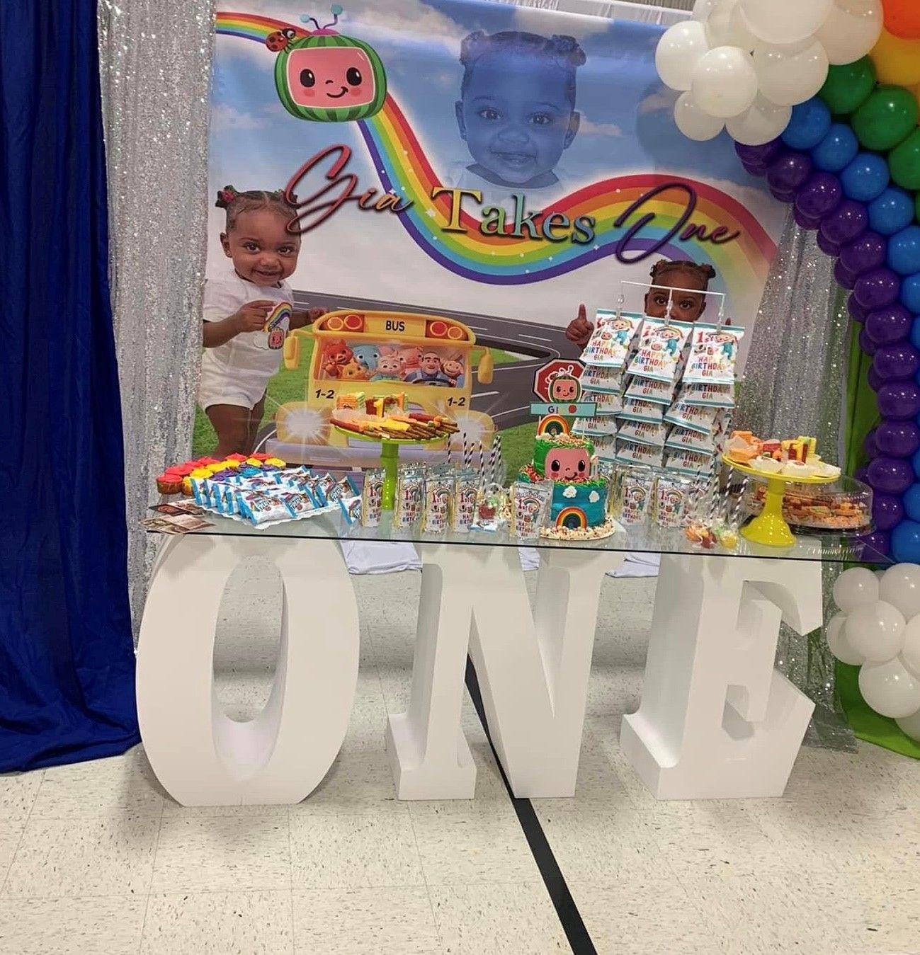 A first birthday party with a Cocomelon theme. A table is decorated with treats and gifts, balloons, and photos.