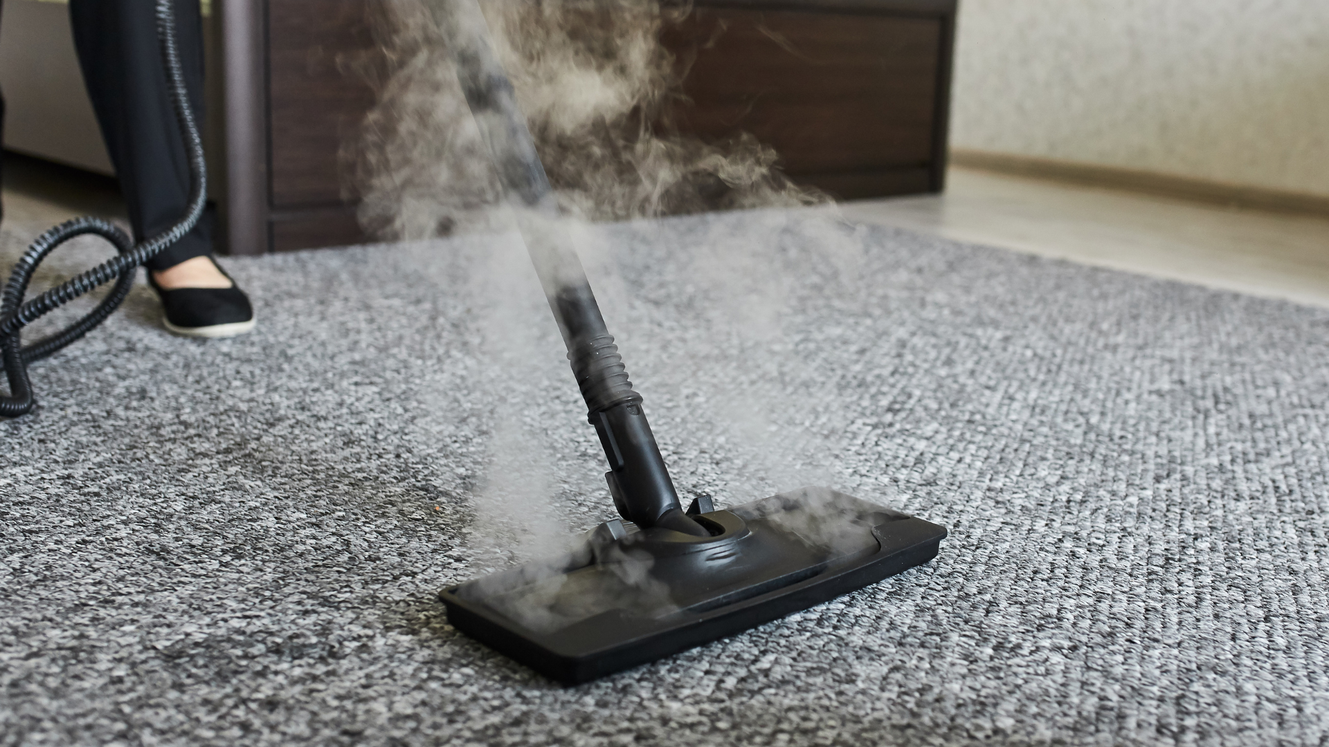 Steam rising from a small black floor steamer on a gray carpet near a person’s foot