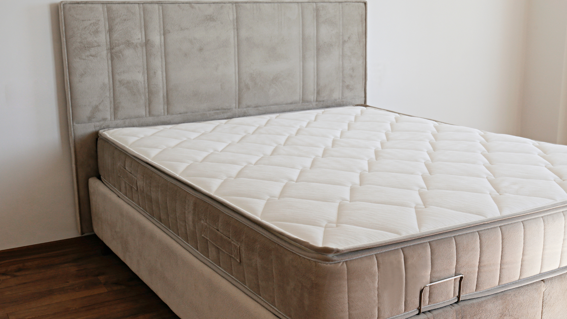 Empty bed with a beige upholstered headboard and white quilted mattress in a simple bedroom