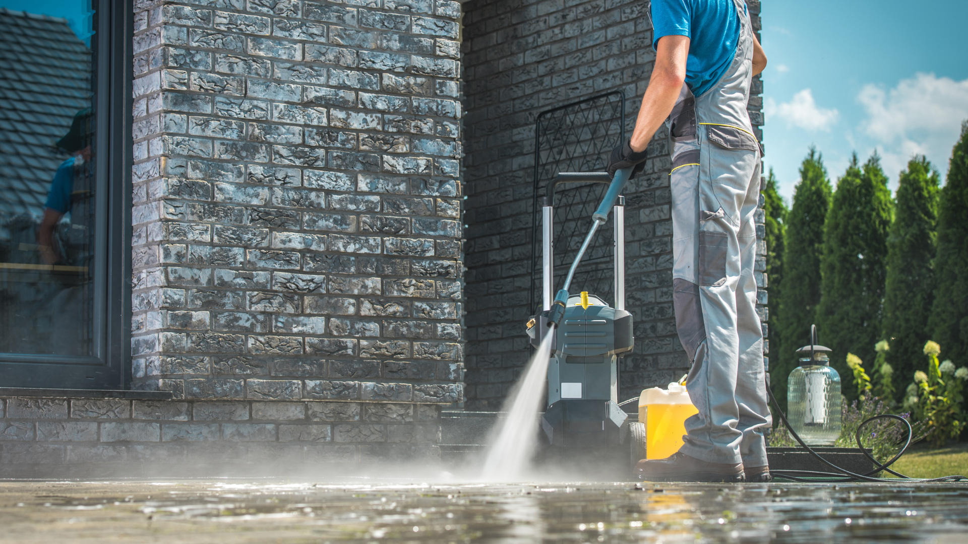 Pressure washing a brick wall and patio beside a house, with water spraying across the wet ground.