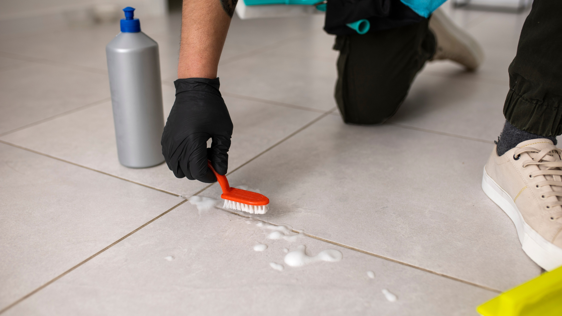 Gloved person scrubbing a tiled floor spill with a brush beside a cleaning bottle