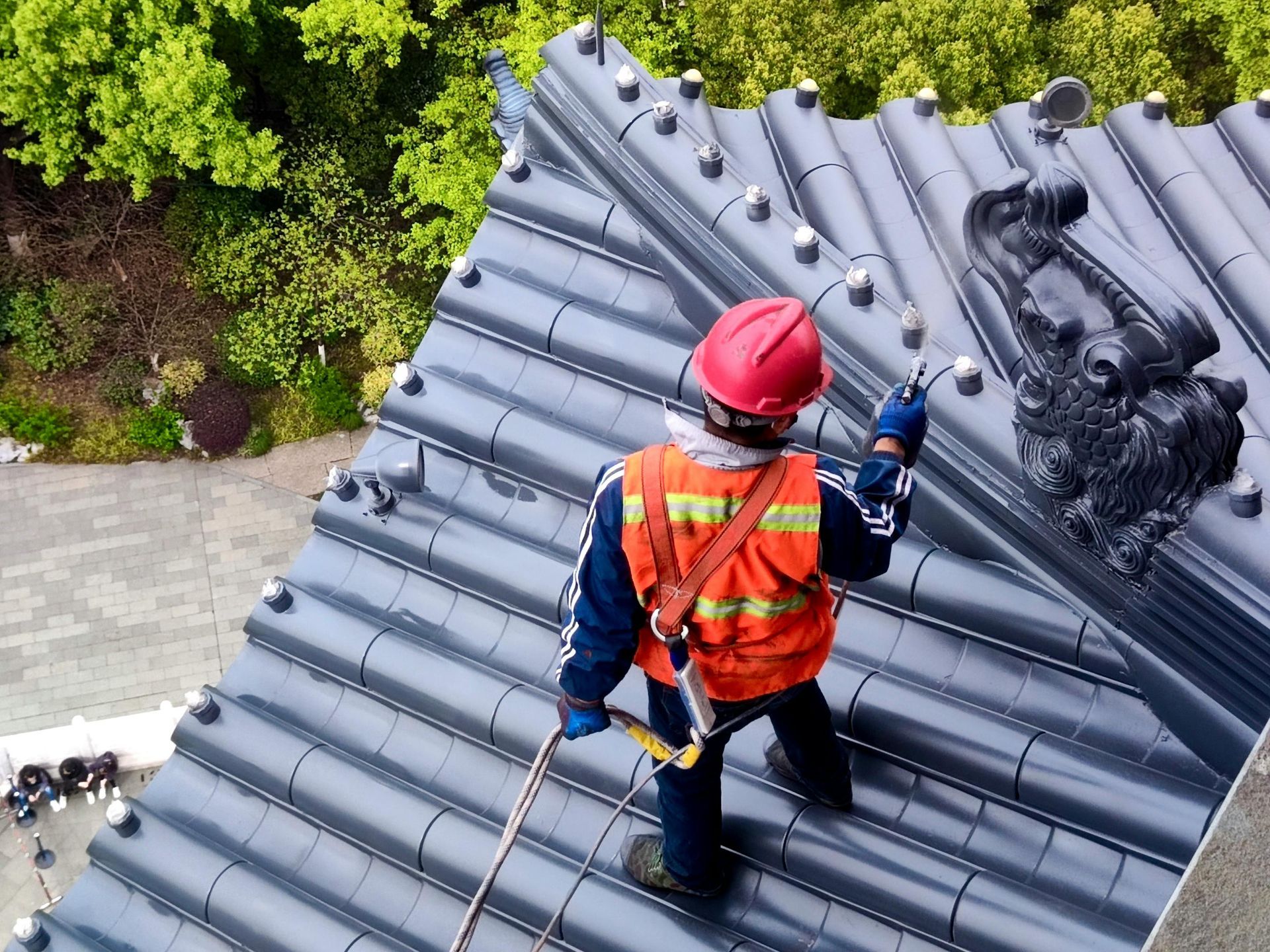 Person on a roof repairing it; wearing safety gear and holding a tool. Green trees in background.