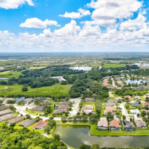 Aerial view of a suburban neighborhood with houses, trees, lakes, and a blue sky with clouds.