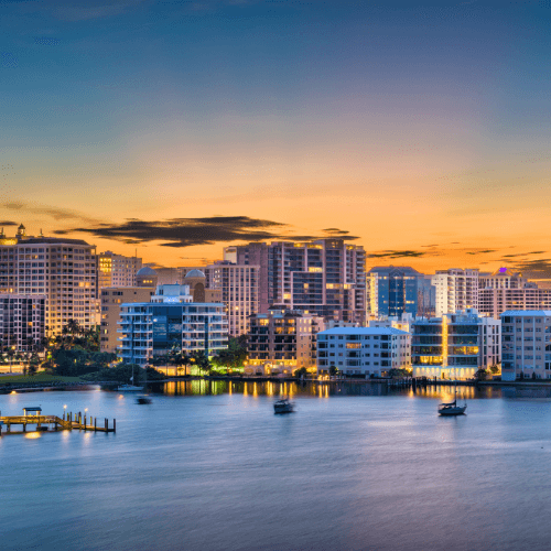 Skyline of West Palm Beach, Florida at dusk with buildings, water, and boats, under a colorful sunset.