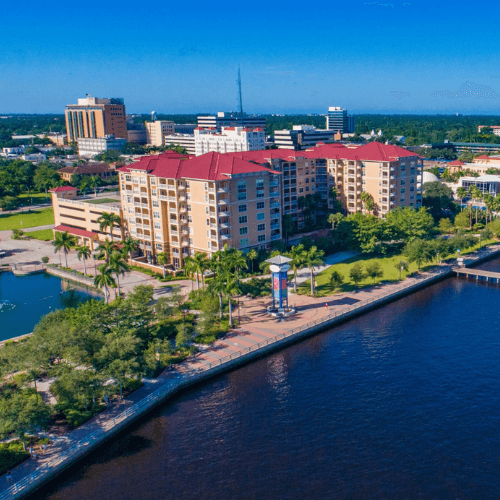 Waterfront buildings with red roofs along a blue river. City skyline in the background, sunny day.