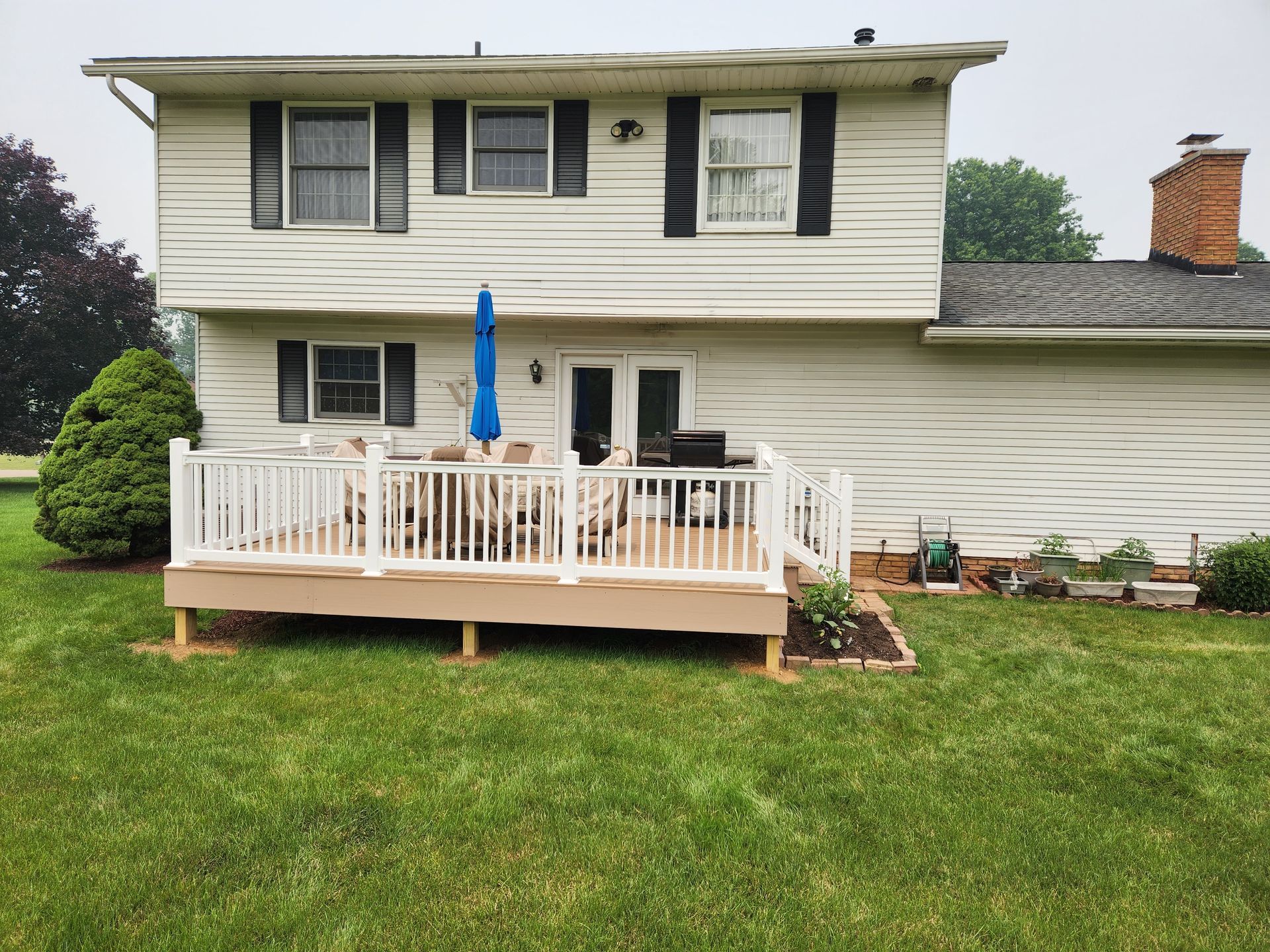 A two-story house with white siding and black shutters, featuring a raised wooden deck with a white railing and umbrella.