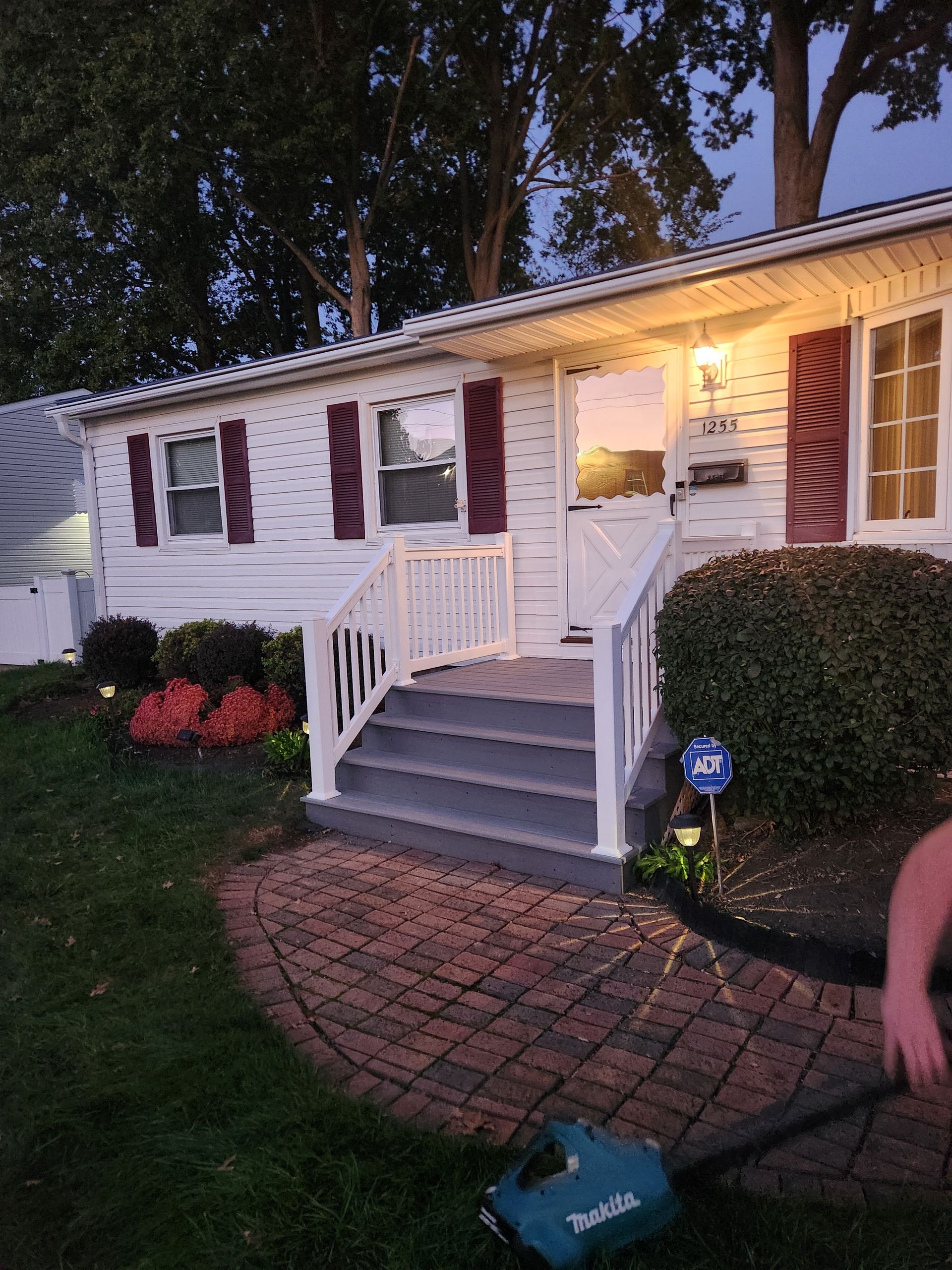 A person using a teal leaf blower on a brick patio in front of a white house with burgundy shutters and a gray staircase.