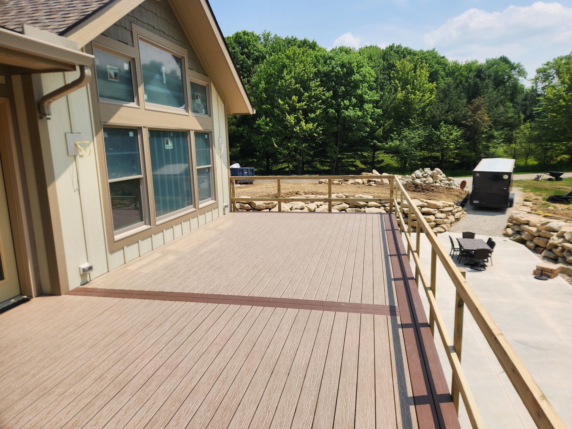 A tan deck with a brown border and wooden railing overlooking a patio, a stone retaining wall, and lush green trees.