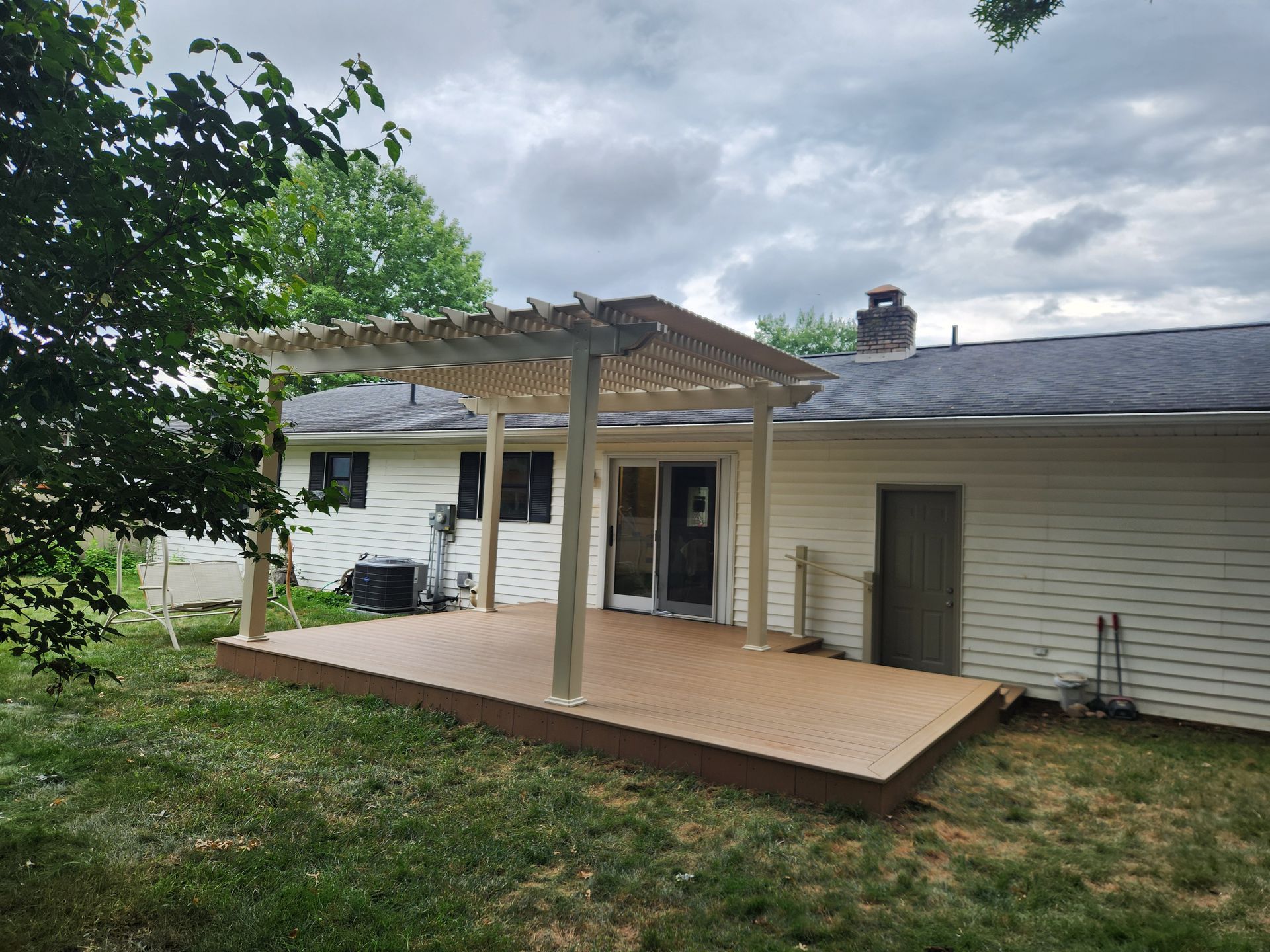 A tan composite deck with a pergola attached to the rear of a white house on a cloudy day.
