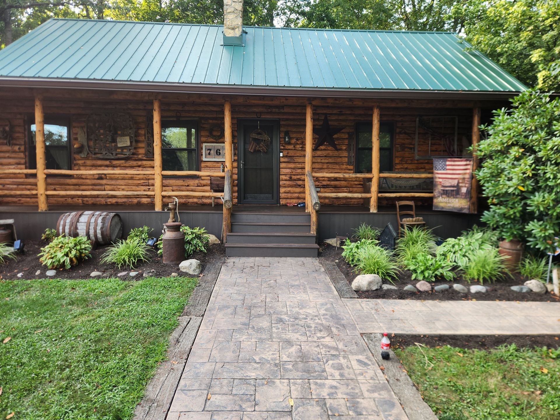 A one-story log cabin with a green metal roof, a front porch, and a stone walkway leading to the entrance.