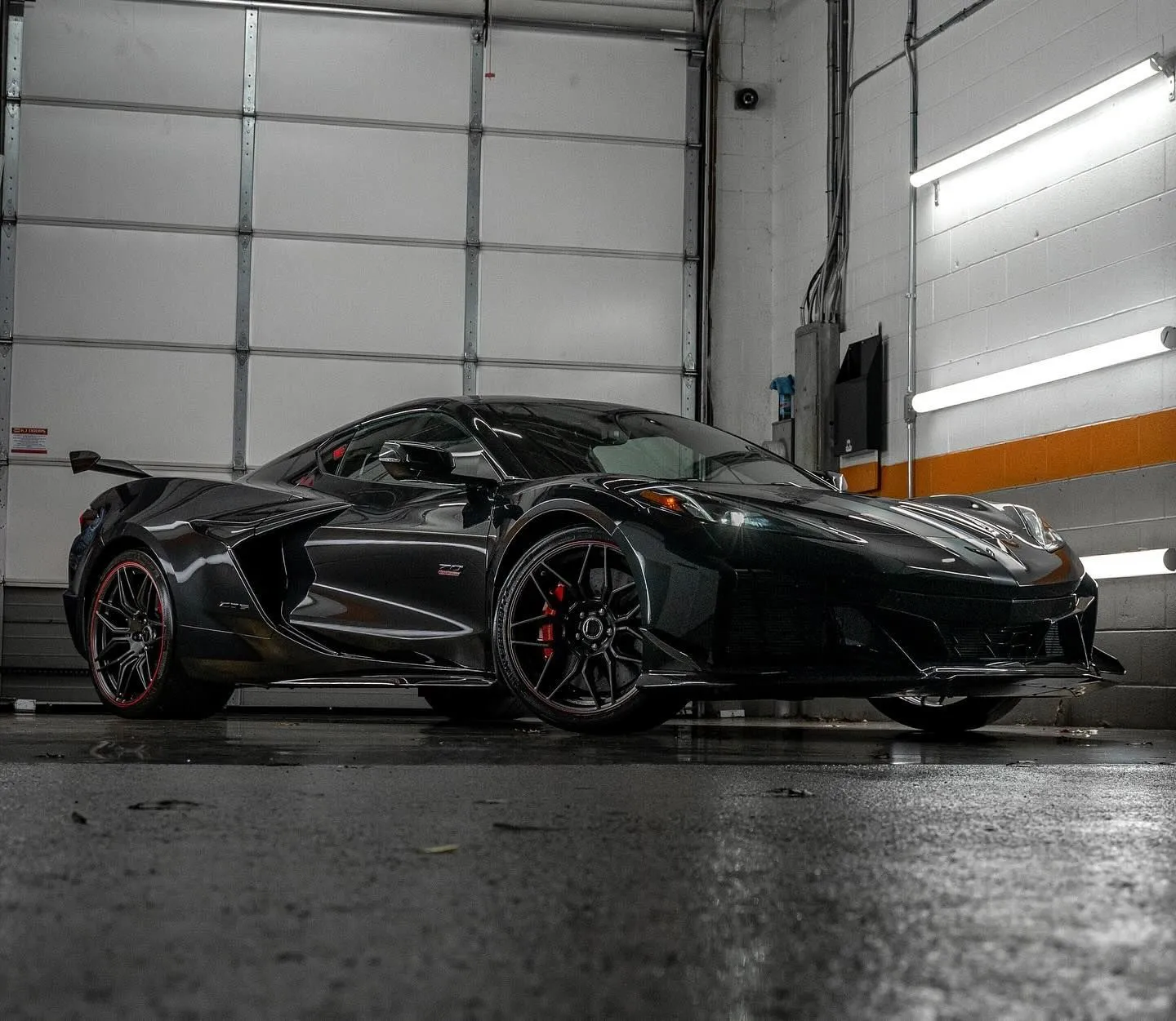 Black sports car in a garage, low angle view with wet floor and bright fluorescent lights.
