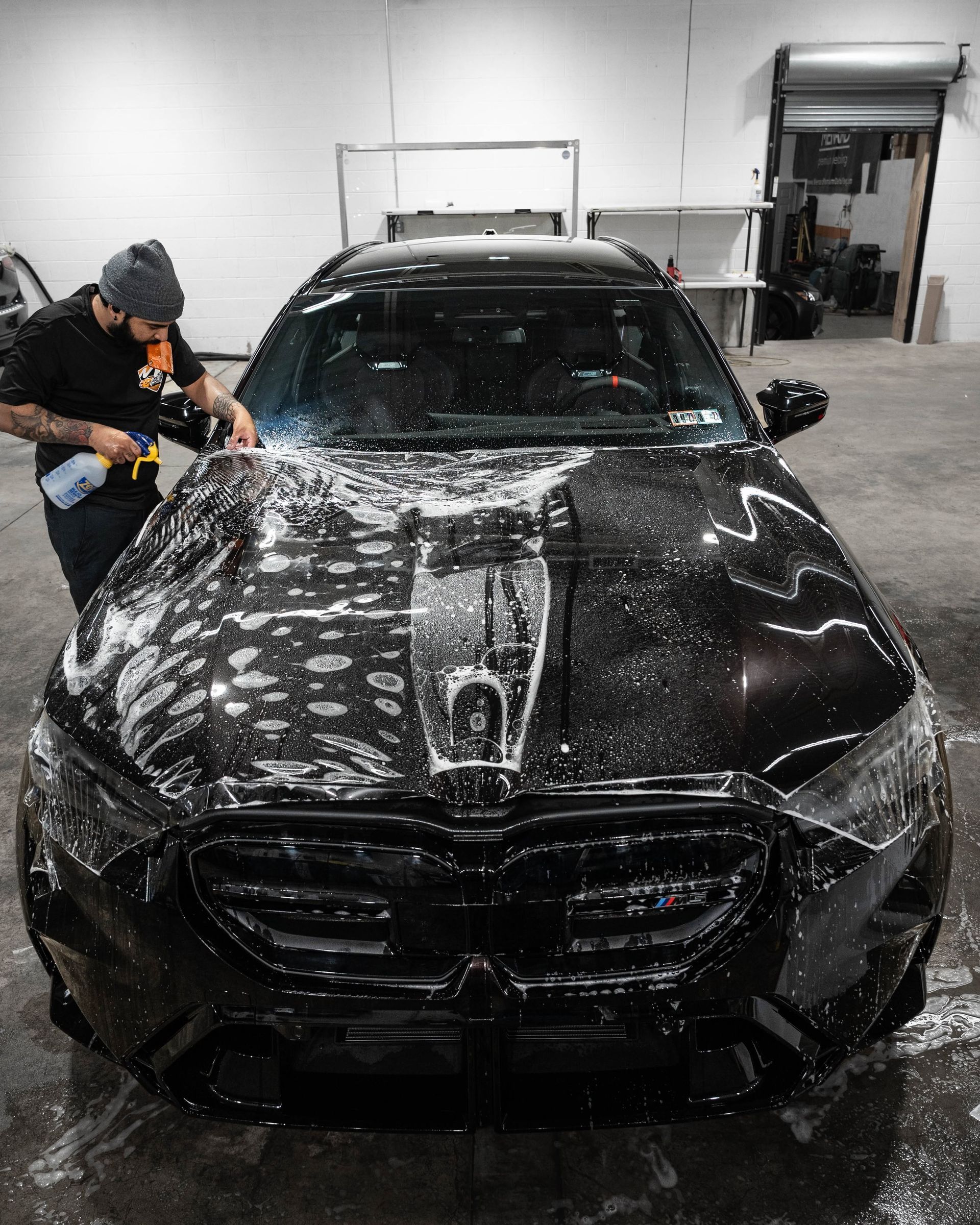 Person washing a black car in a garage with soap suds and water spray