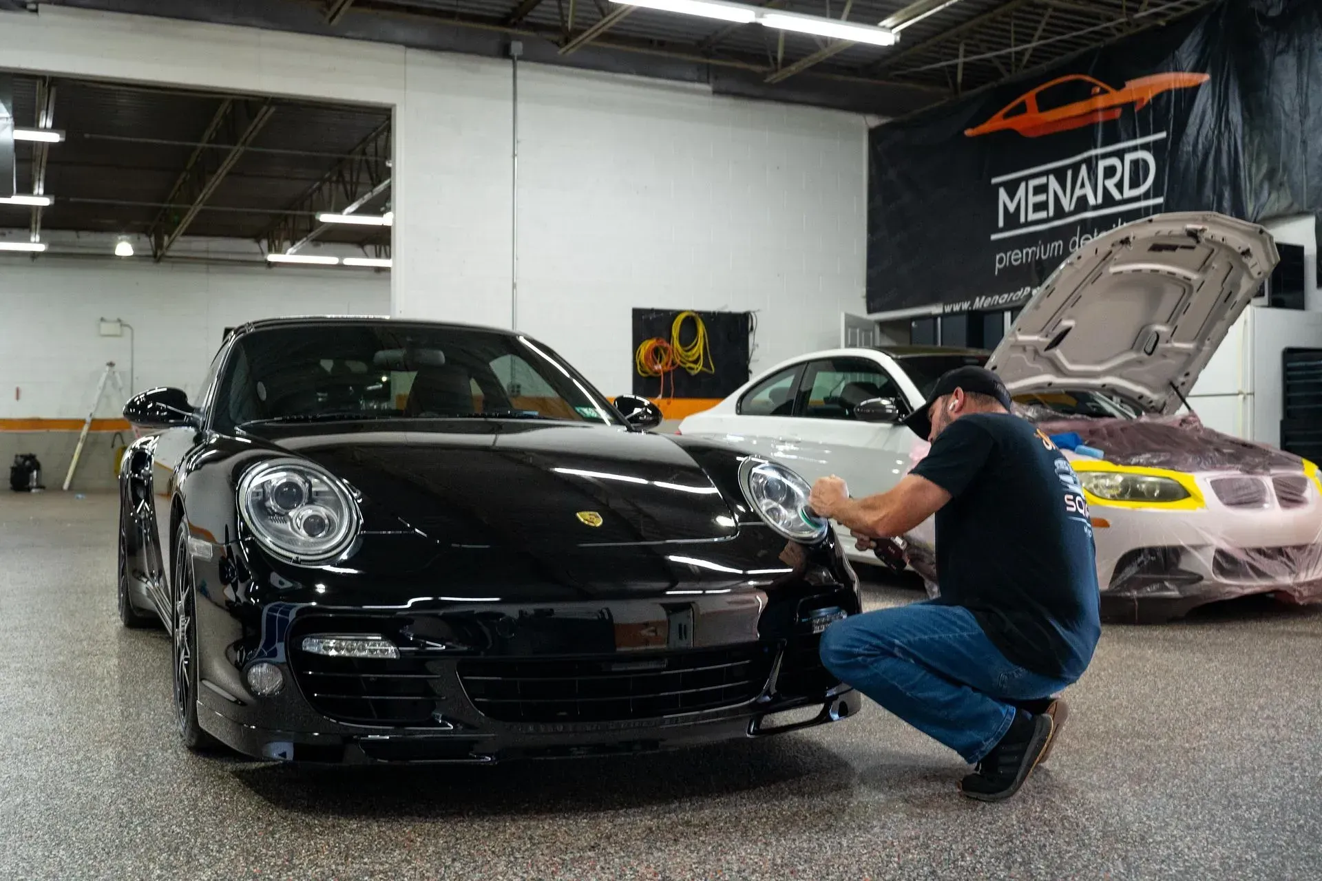 Man polishing a black Porsche in a showroom, with a white car and Menard sign in the background