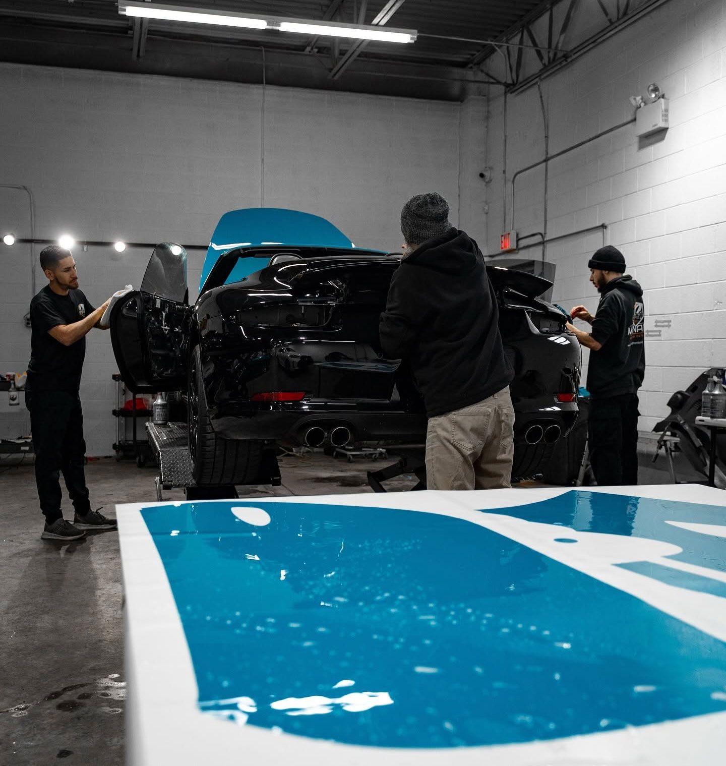 People working on a black car in a garage, with a blue vehicle panel in the foreground.