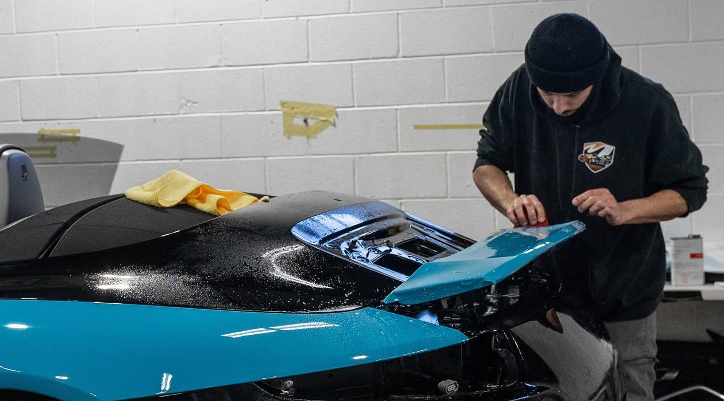 Person working on a blue sports car in a garage, with a raised rear spoiler and tools nearby.