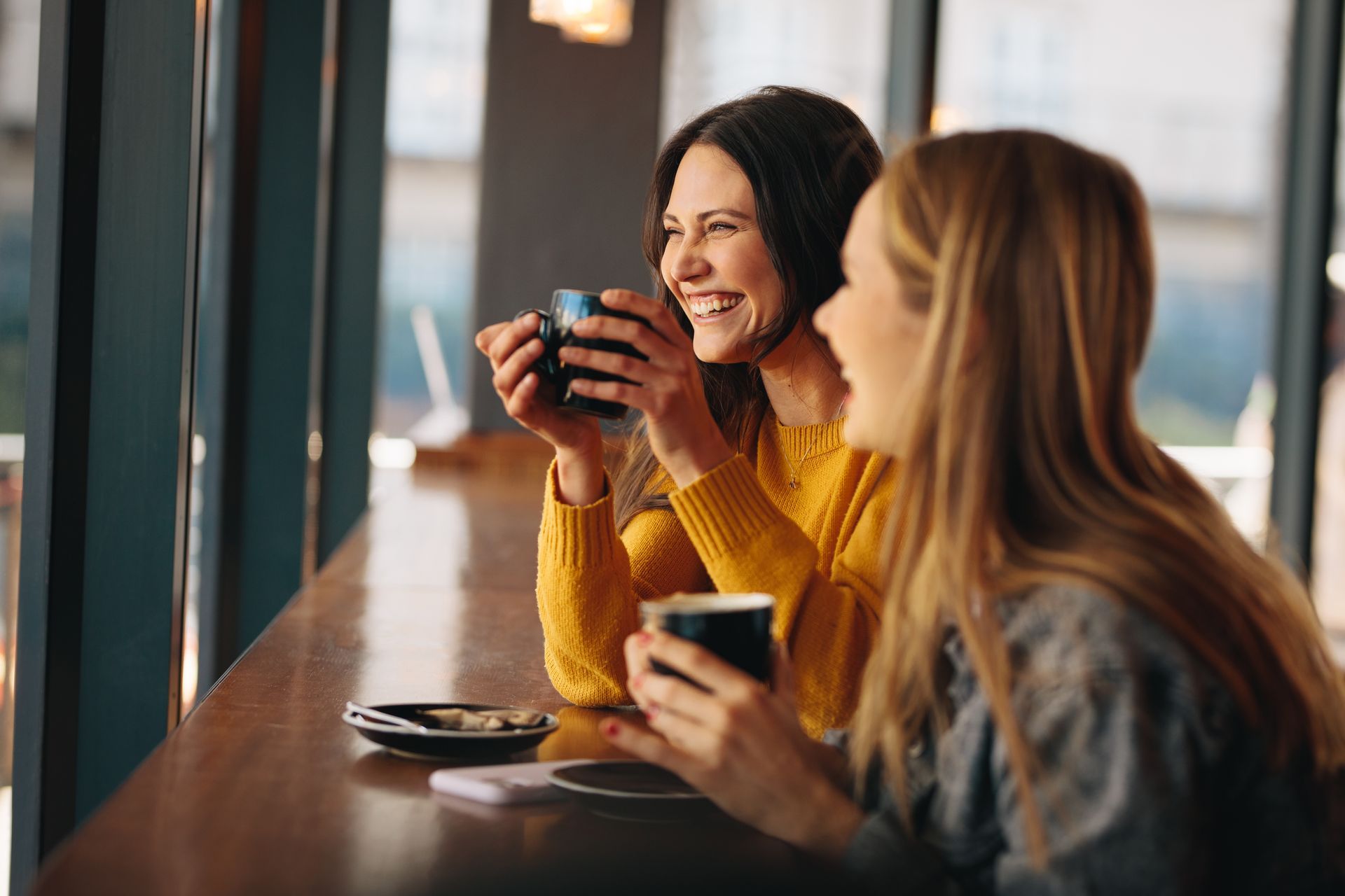 Two women in a cafe, smiling and holding coffee cups, seated by a window.