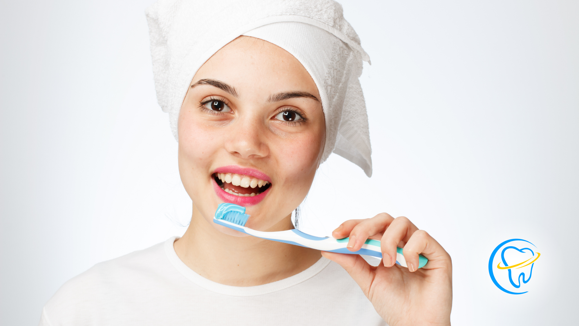 Woman with towel on head, holding a toothbrush with toothpaste, smiling.