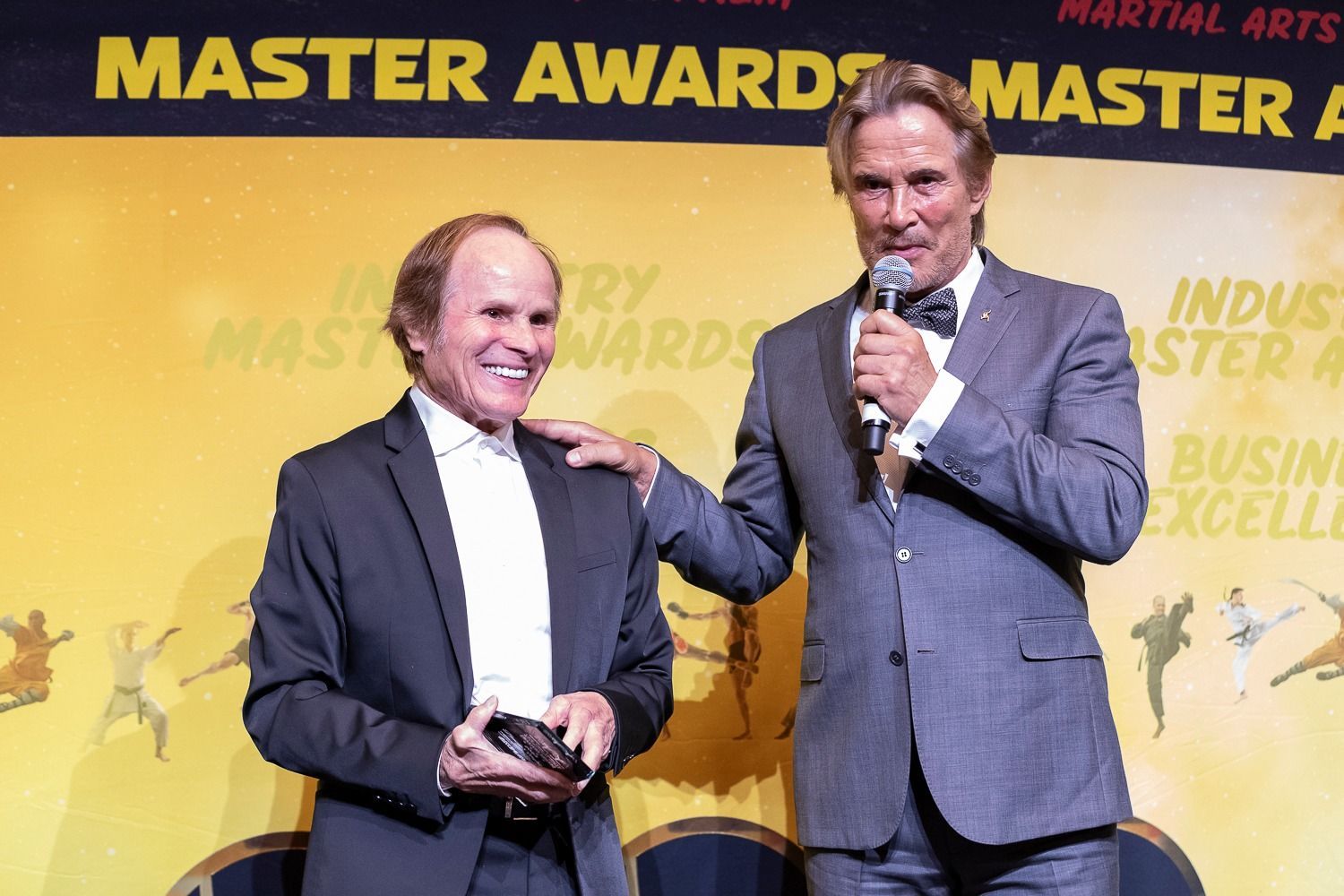 Two men at an awards ceremony. One in a suit smiles, the other speaks into a mic, patting his shoulder. Yellow background.