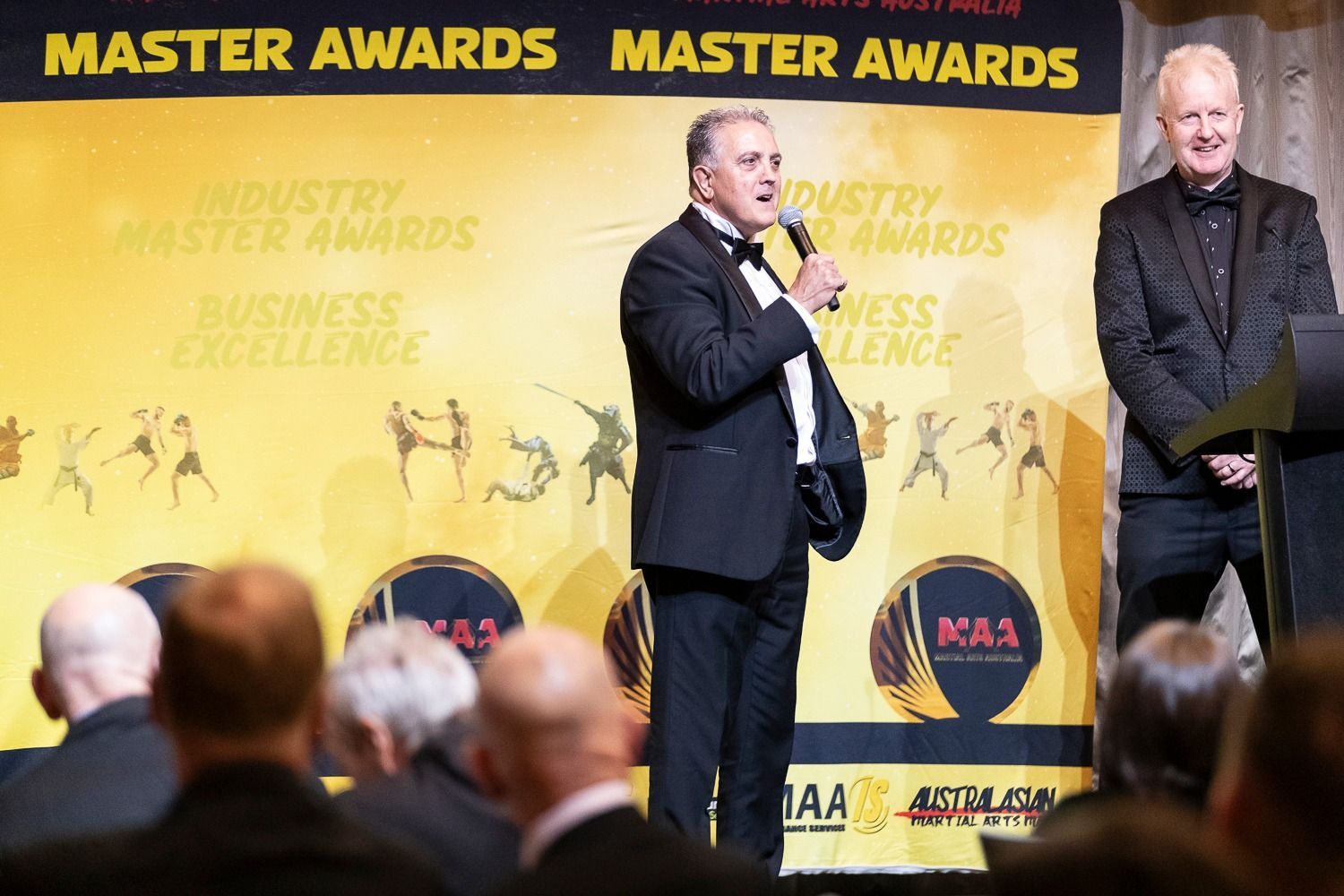 Two men at a formal awards ceremony. One speaks into a microphone, another stands by. Yellow background.
