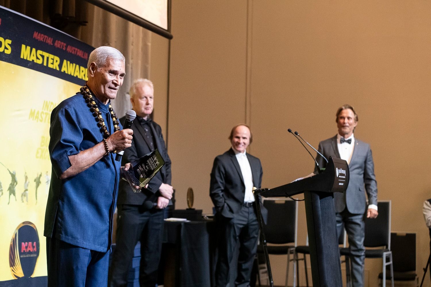 Men on stage at an awards ceremony, one speaking into a microphone, others holding trophies.