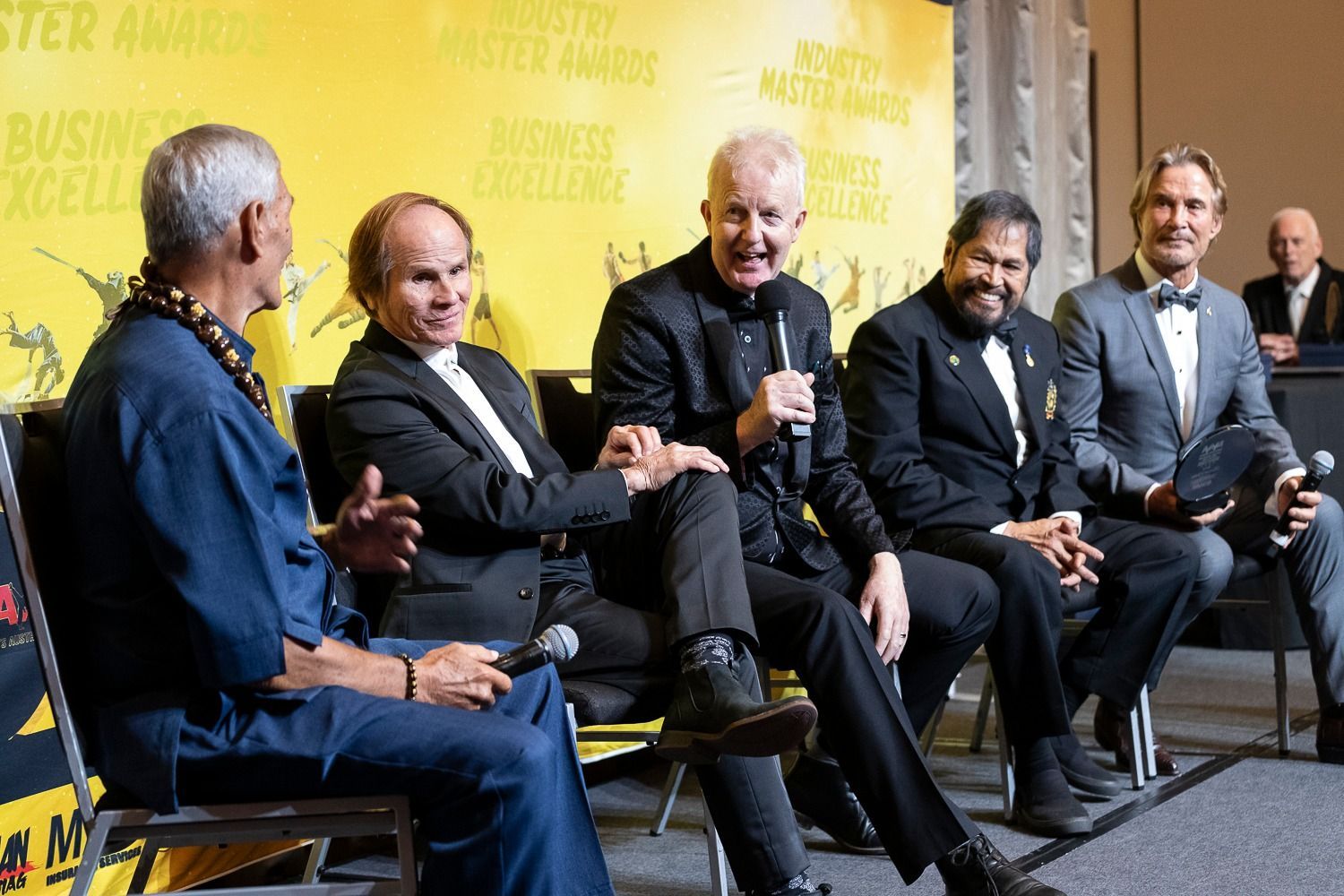 Five men seated, speaking at an event. Backdrop with awards text. The man in the center speaks into a microphone.