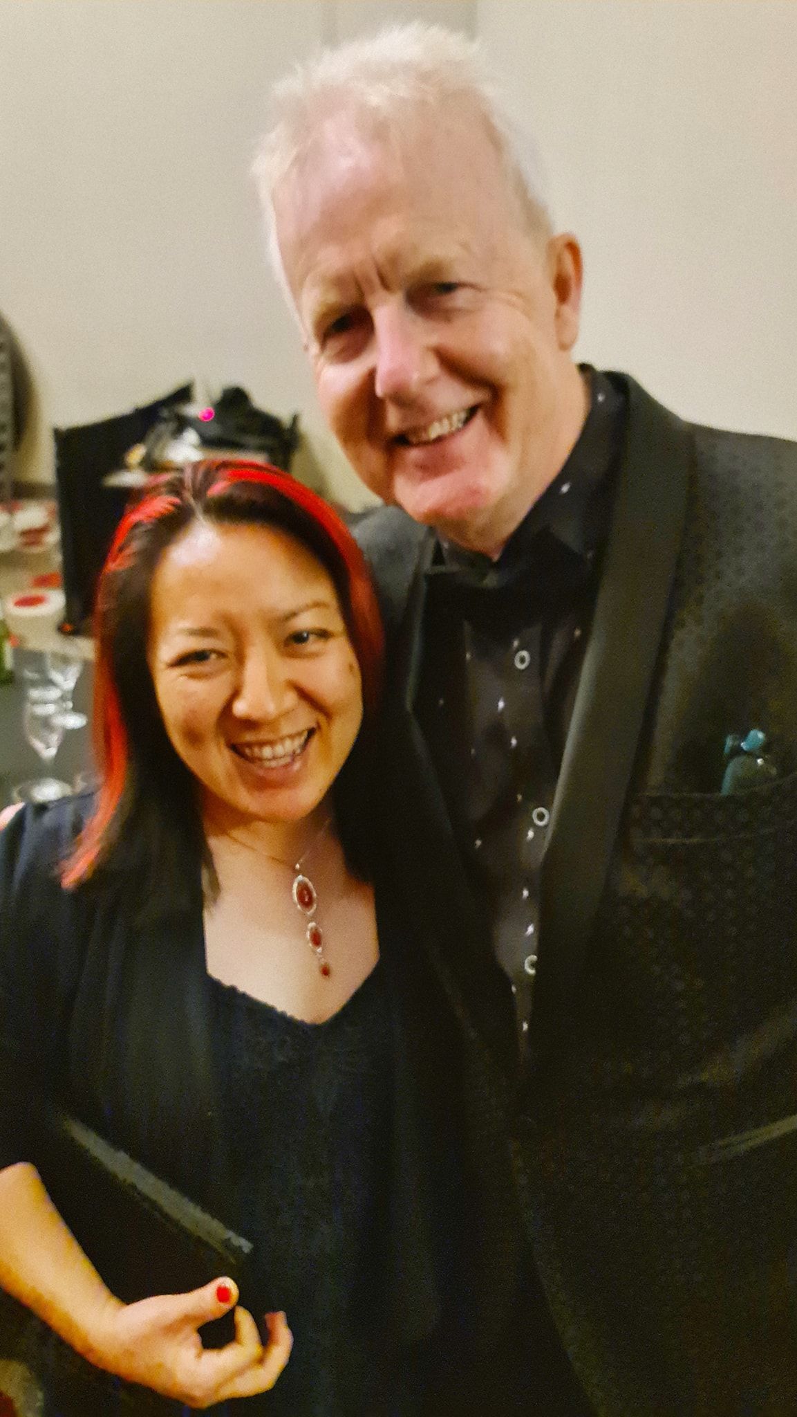 Woman with red-streaked hair and smiling man pose together. Man wears a patterned shirt and a jacket. Both smile.