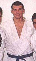 A man in a white karate uniform with a blue belt is standing next to two other men.