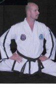A man in a white karate uniform with a black belt is sitting on the floor.