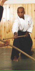 A man in a white kimono is kneeling down holding two wooden sticks.