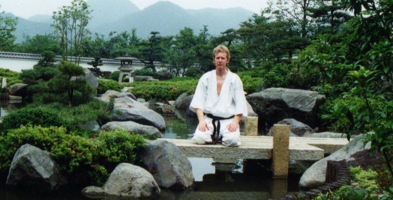A man in a karate uniform is sitting on a bridge over a pond.