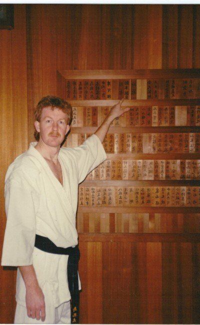 A man in a white robe and black belt stands in front of a wooden wall