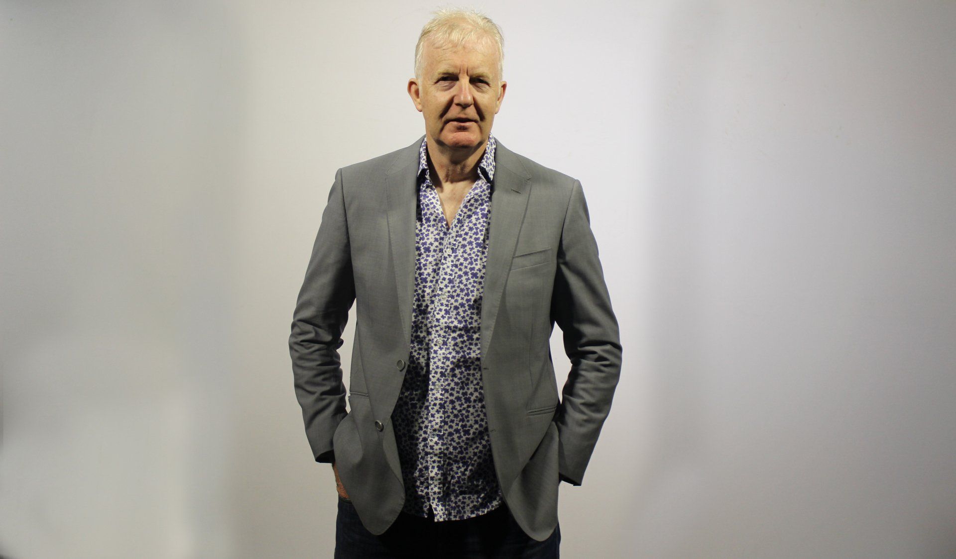 A man in a suit and a floral shirt is standing with his hands in his pockets.