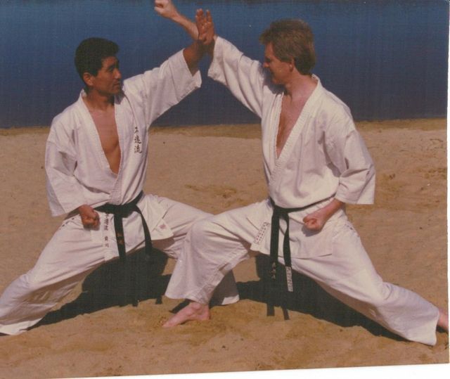 Two men in white karate uniforms kneeling on the beach