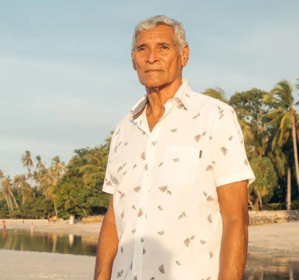 Older man in white shirt on a beach, looking at the camera, with trees and water in the background.