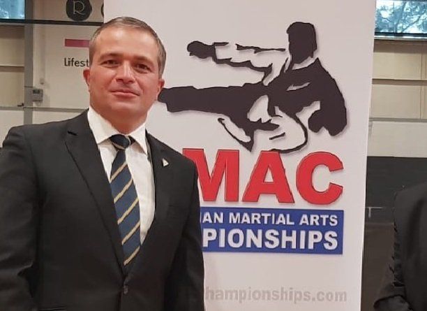 A man in a suit and tie stands in front of a sign that says mac an martial arts pionships