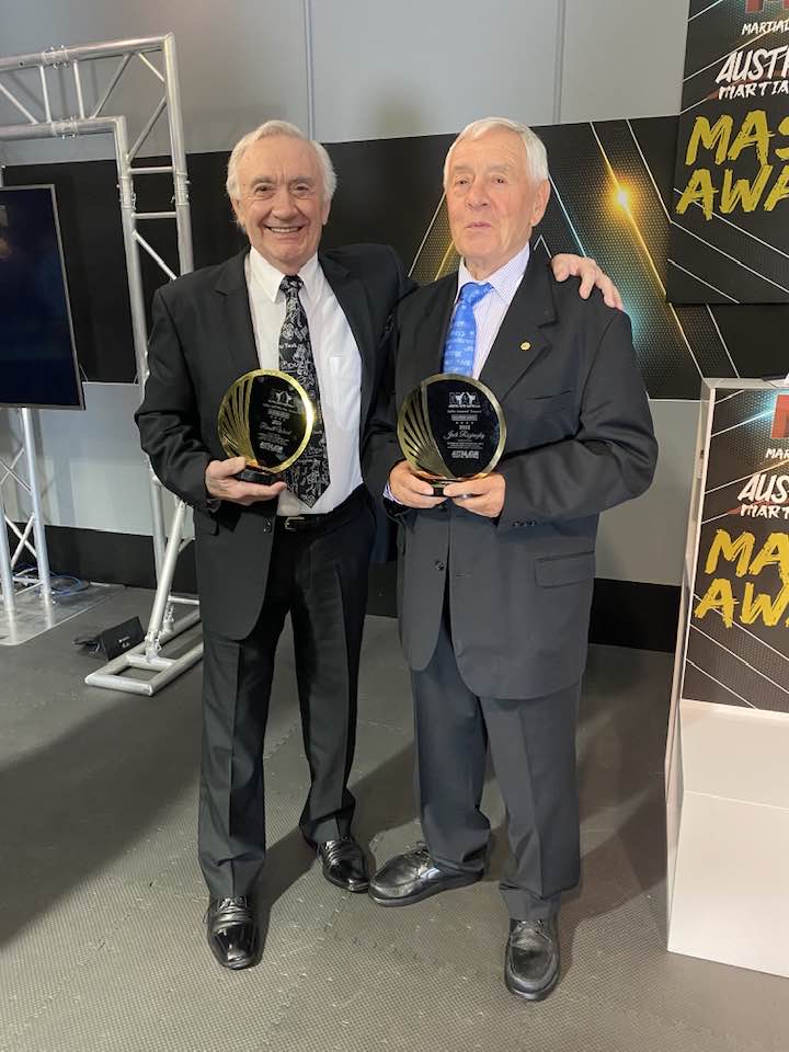 Two men in suits and ties are standing next to each other holding trophies.