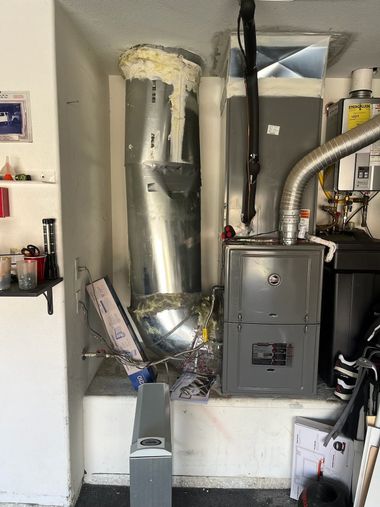 Furnace and ductwork in a garage, gray metal with white insulation. Various pipes and a water heater visible.
