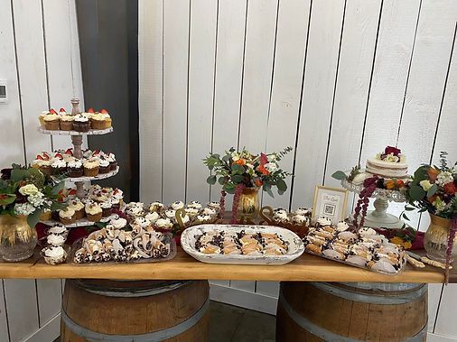 Dessert display on a wooden table with cupcakes, pastries, and floral centerpieces against a white fence backdrop