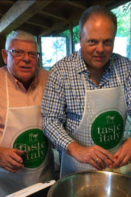 Two men in aprons stirring food in a large pot at a cooking demo, one smiling at the camera.