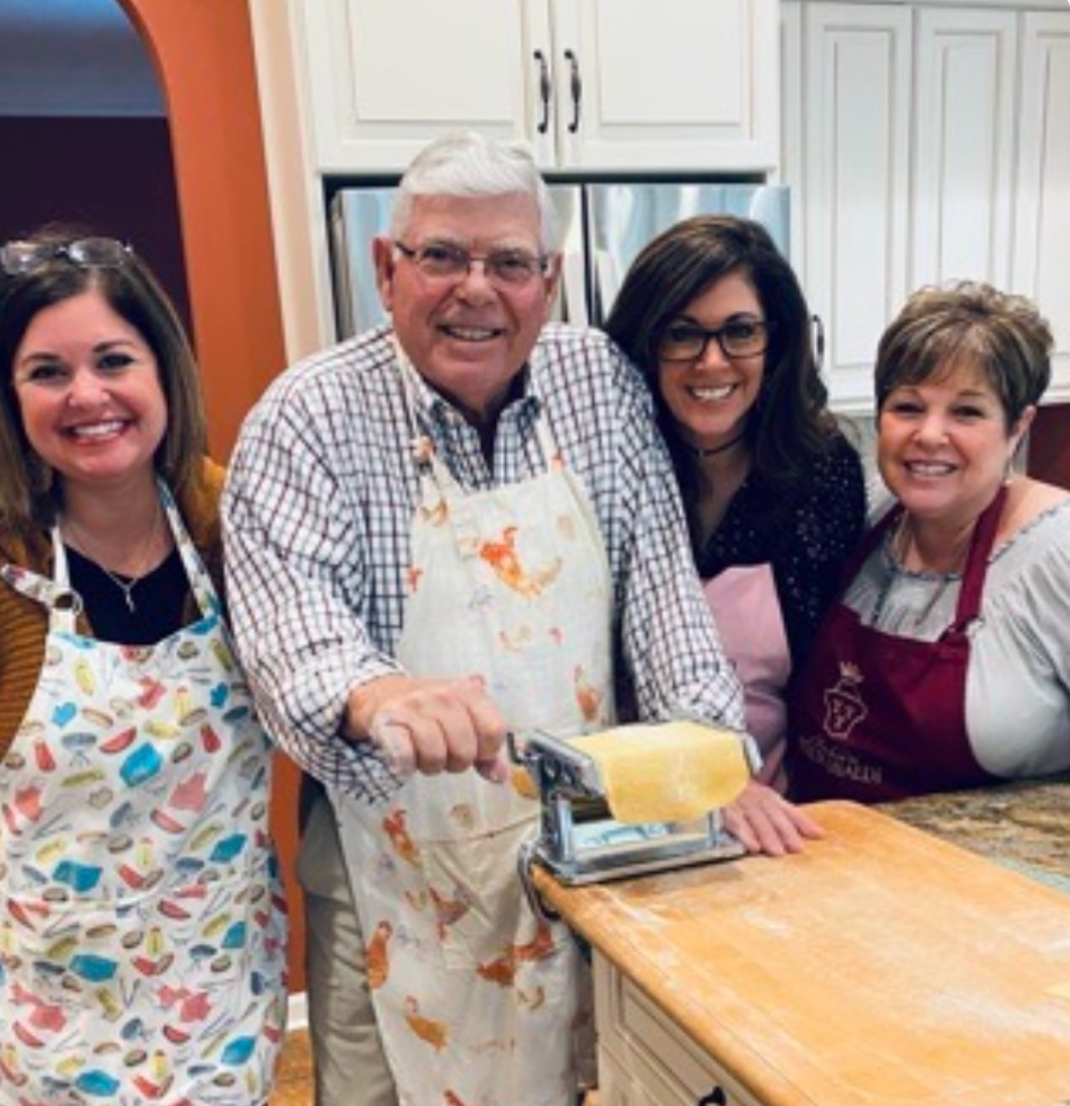 Four people in aprons pose in a kitchen, smiling beside a pasta roller and countertop dough.