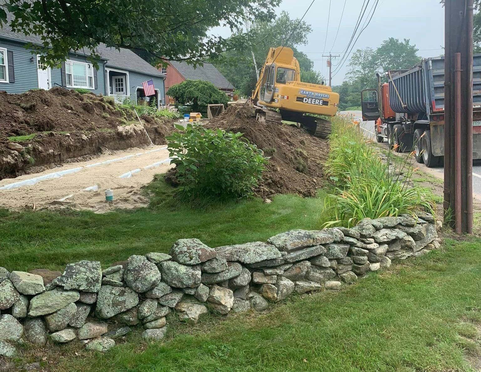 A large yellow excavator is sitting on top of a pile of dirt next to a stone wall.