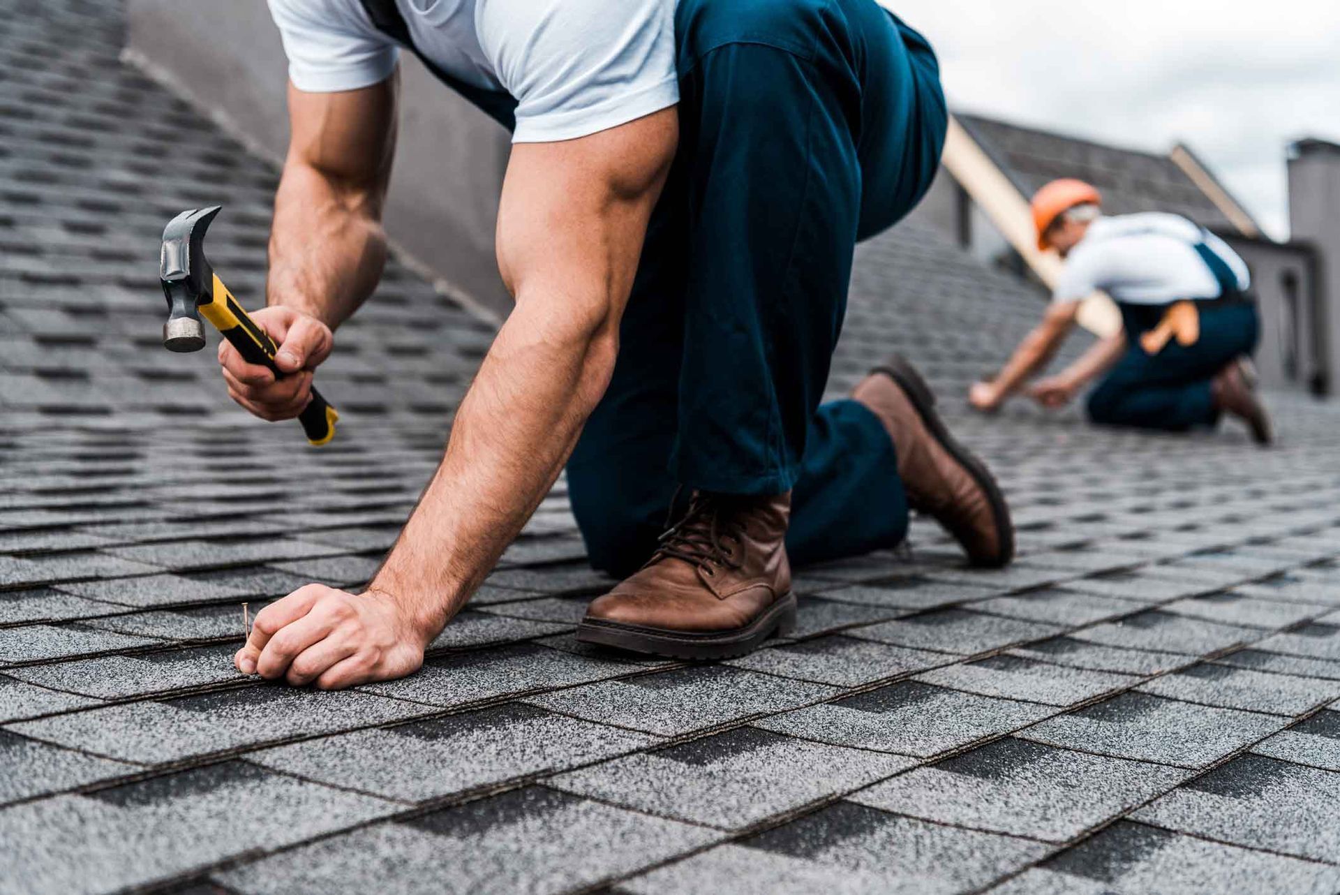 Workers installing asphalt shingles on a roof, one kneeling with a hammer in the foreground.