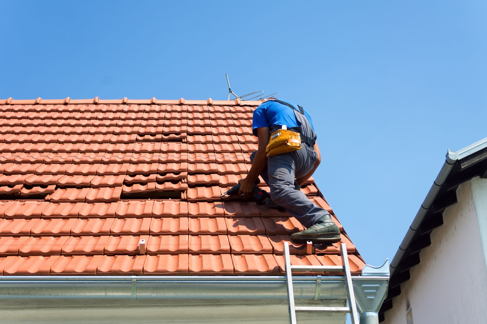 A Worker on a ladder repairing red roof tiles under clear blue sky.