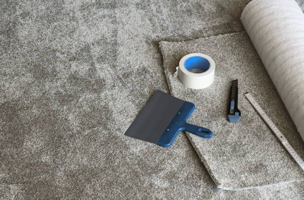 A roll of carpet , a spatula , tape , and a cutter are on the floor.