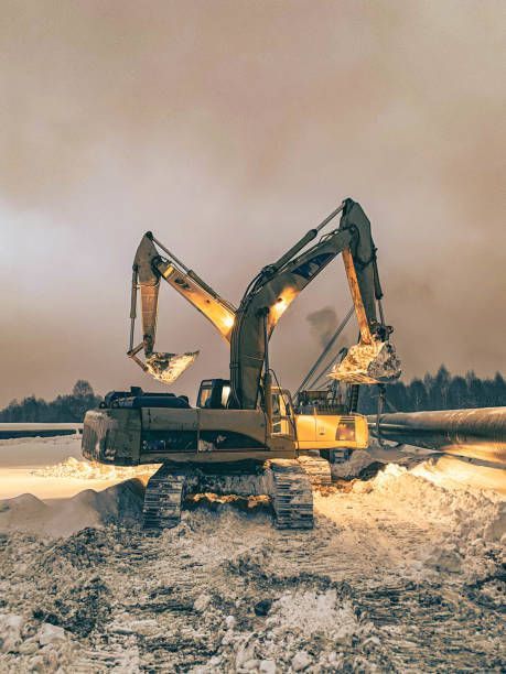 Two excavators working, illuminated by lights, in a snowy landscape.