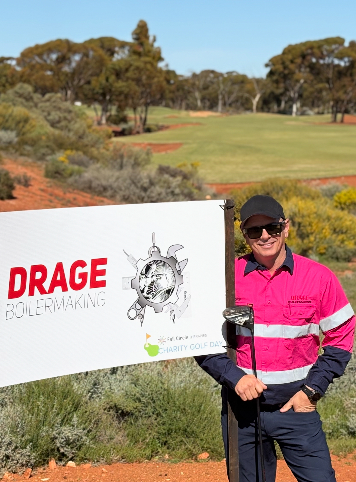A person in a bright pink and navy high-vis shirt stands next to a Drage Boilermaking sign on a sunny golf course.