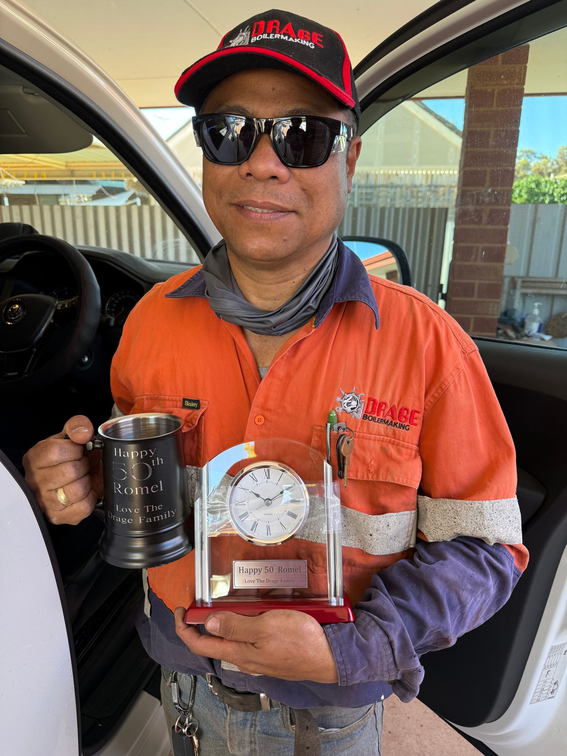 A person in a high-visibility orange shirt and cap sits in a vehicle, holding a beer mug and a glass desktop clock.